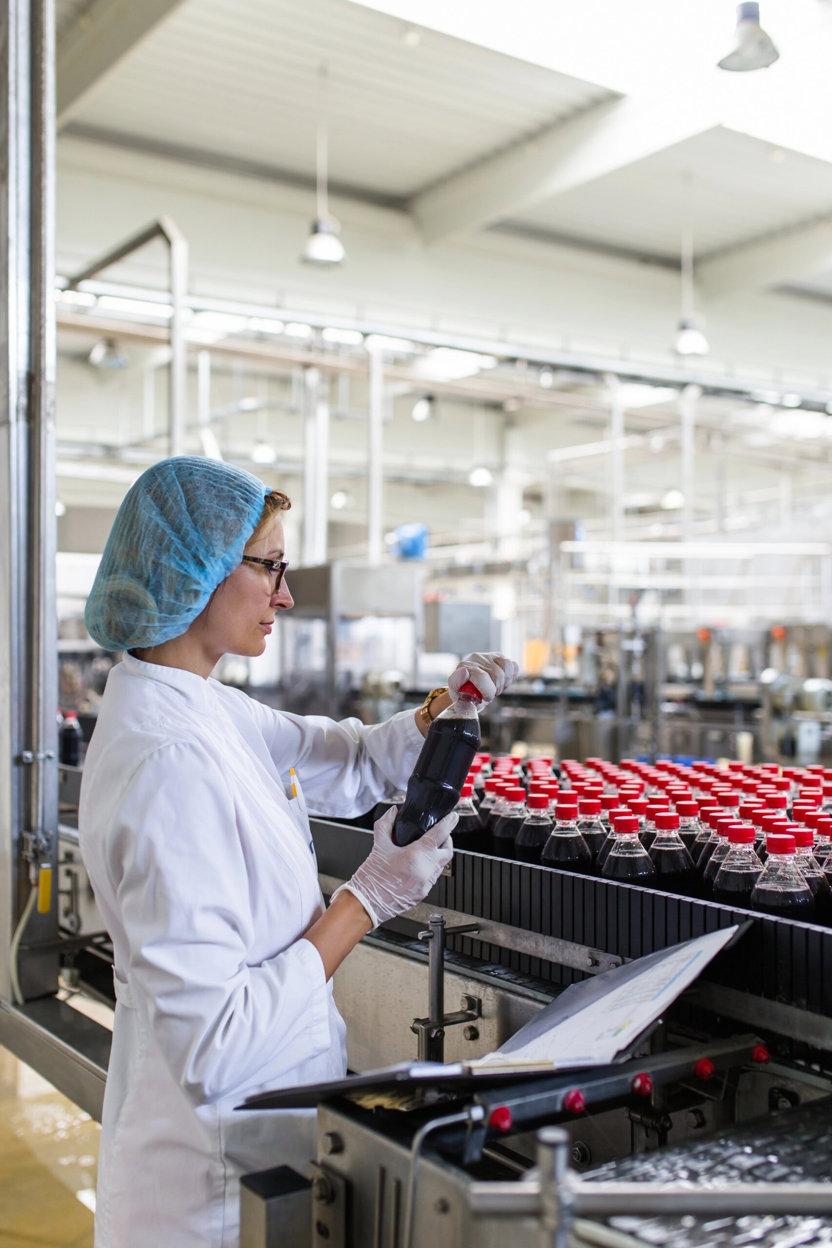 Serious middle age woman quality control worker checking robotic line for bottling and packaging carbonated black juice of soft drink into bottles.